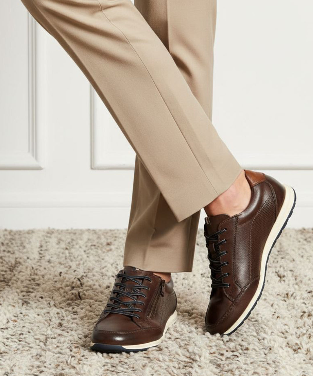 Lifestyle shot of bugatti Thorello brown premium leather sneakers with white soles, styled with tan trousers on a carpet.