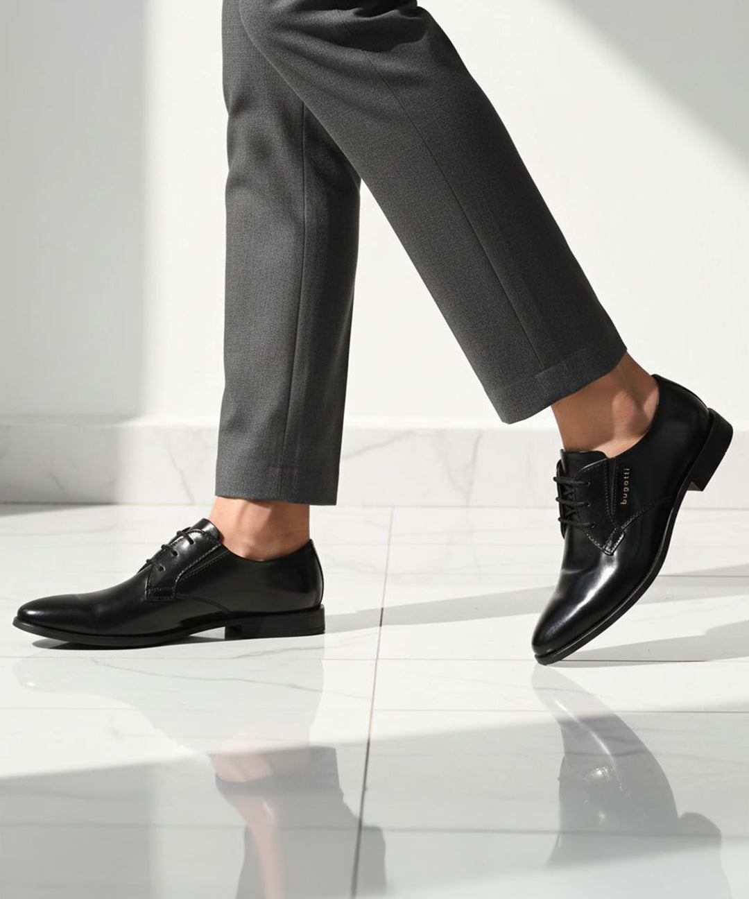 Side view of black bugatti leather derby shoes, paired with gray dress pants on a white marble floor.