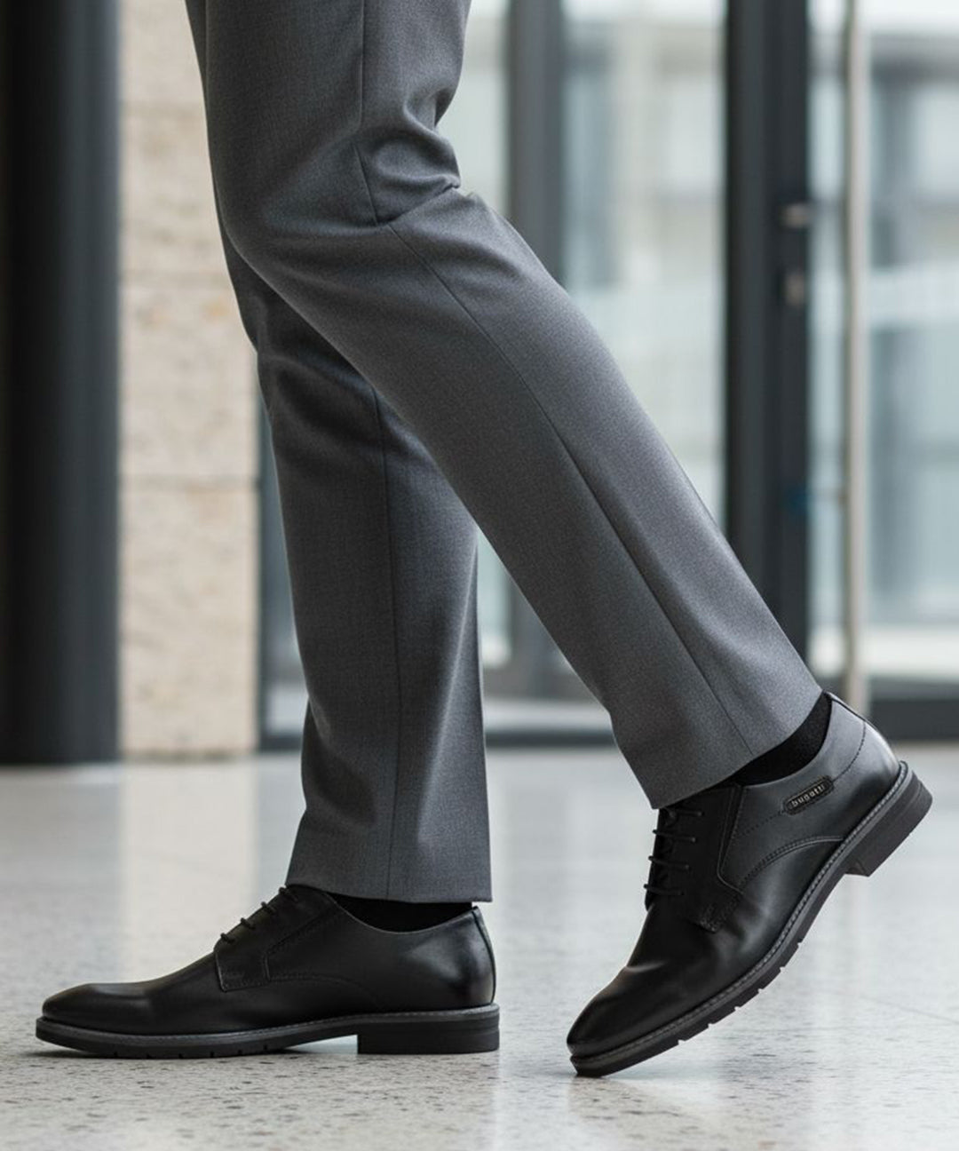 Lifestyle close-up of black bugatti Mero formal leather derby shoes, gray dress pants, and black socks.
