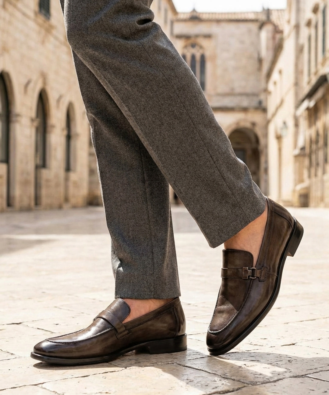 Lifestyle shot of bugatti dark green leather penny loafers with grey dress pants on a stone tiled street.
