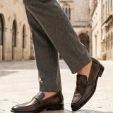 Lifestyle shot of bugatti dark green leather penny loafers with grey dress pants on a stone tiled street.
