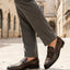 Lifestyle shot of bugatti dark green leather penny loafers with grey dress pants on a stone tiled street.

