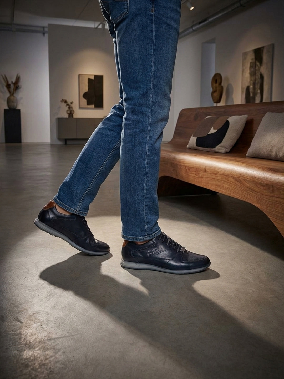 Lifestyle view of bugatti Thorello dark blue leather sneakers with blue jeans and casual indoor setting.
