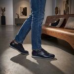 Lifestyle view of bugatti Thorello dark blue leather sneakers with blue jeans and casual indoor setting.
