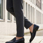 Cropped lifestyle shot of bugatti Rico black leather slip-on formal shoes, paired with gray dress pants.

