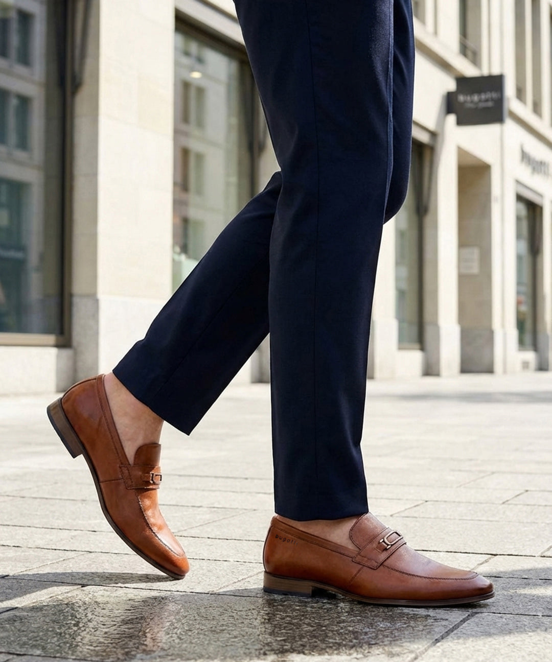 Cropped lifestyle image showing bugatti Matina cognac leather slip-on formal shoes with dark trousers.