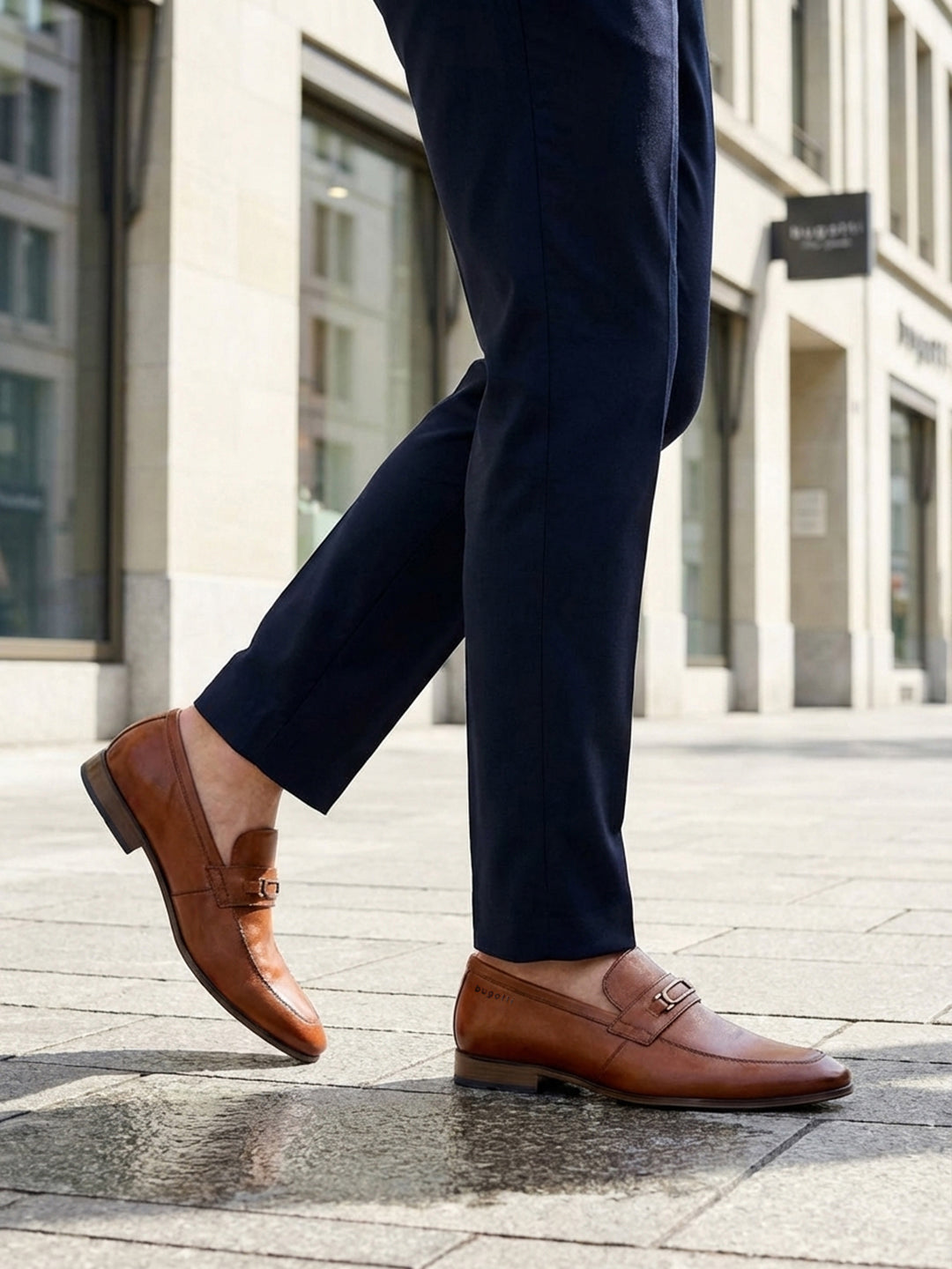 Cropped lifestyle image showing bugatti Matina cognac leather slip-on formal shoes with dark trousers.