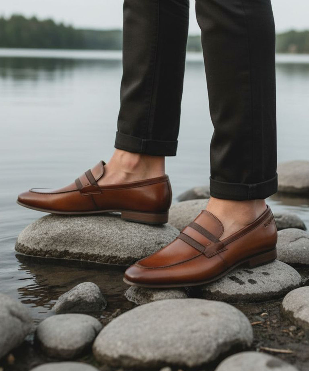 bugatti cognac leather loafers on rocks, paired with dark cuffed pants in a lifestyle shot.
