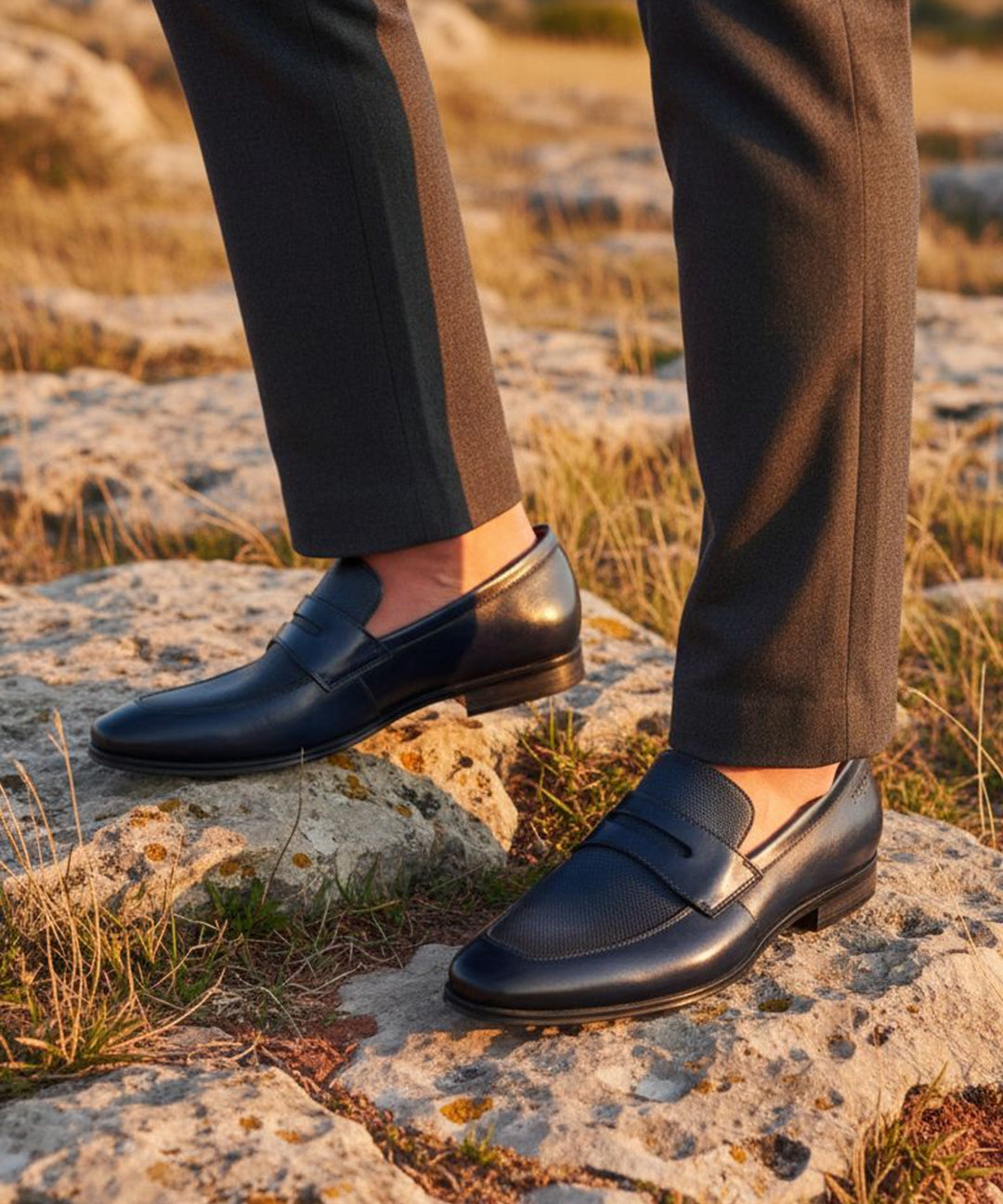 bugatti Margo dark blue premium leather penny loafers worn with gray dress pants on a rocky surface.
