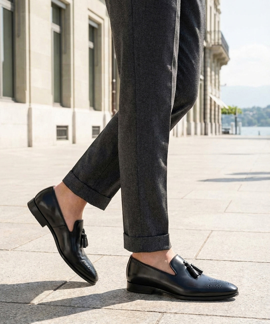 bugatti Margo black leather slip-on shoe with tassel detail, paired with gray trousers.
