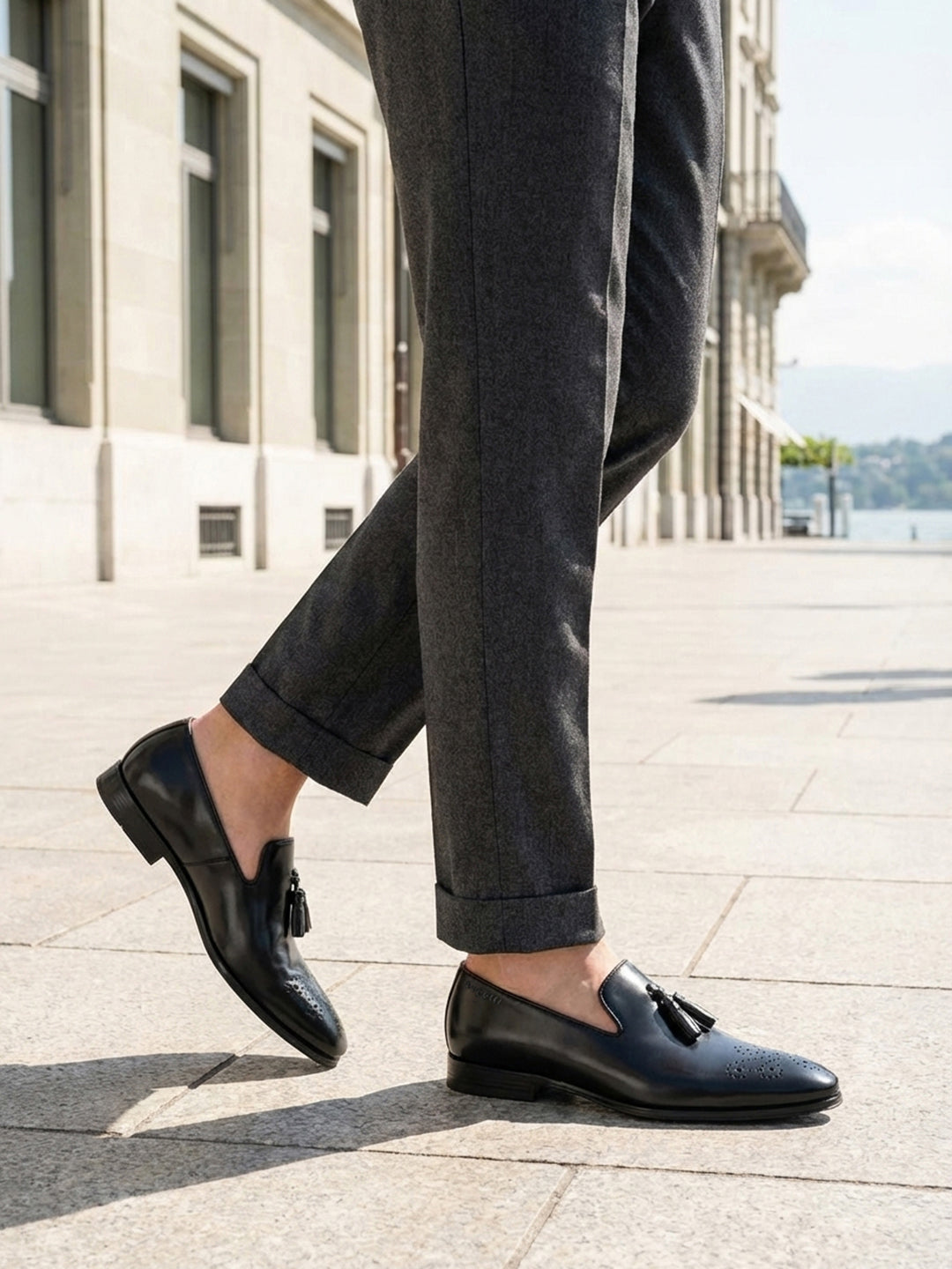 bugatti Margo black leather slip-on shoe with tassel detail, paired with gray trousers.
