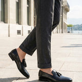 bugatti Margo black leather slip-on shoe with tassel detail, paired with gray trousers.

