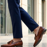bugatti Margo cognac leather slip-on shoes with tassels, worn with dark blue pants in a lifestyle shot.
