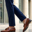 bugatti Margo cognac leather slip-on shoes with tassels, worn with dark blue pants in a lifestyle shot.
