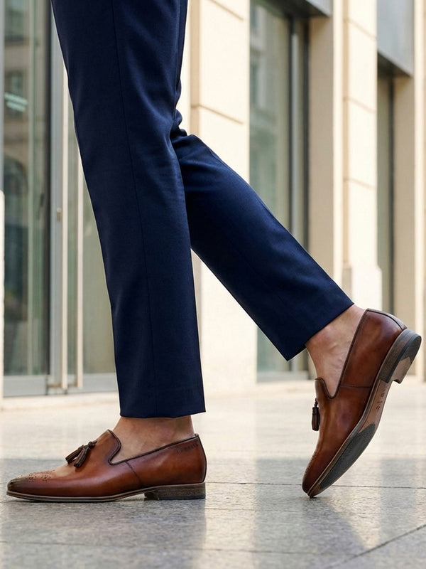bugatti Margo cognac leather slip-on shoes with tassels, worn with dark blue pants in a lifestyle shot.
