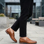 bugatti Sula Revo cognac penny loafers in premium leather, paired with black textured pants, outdoor setting.
