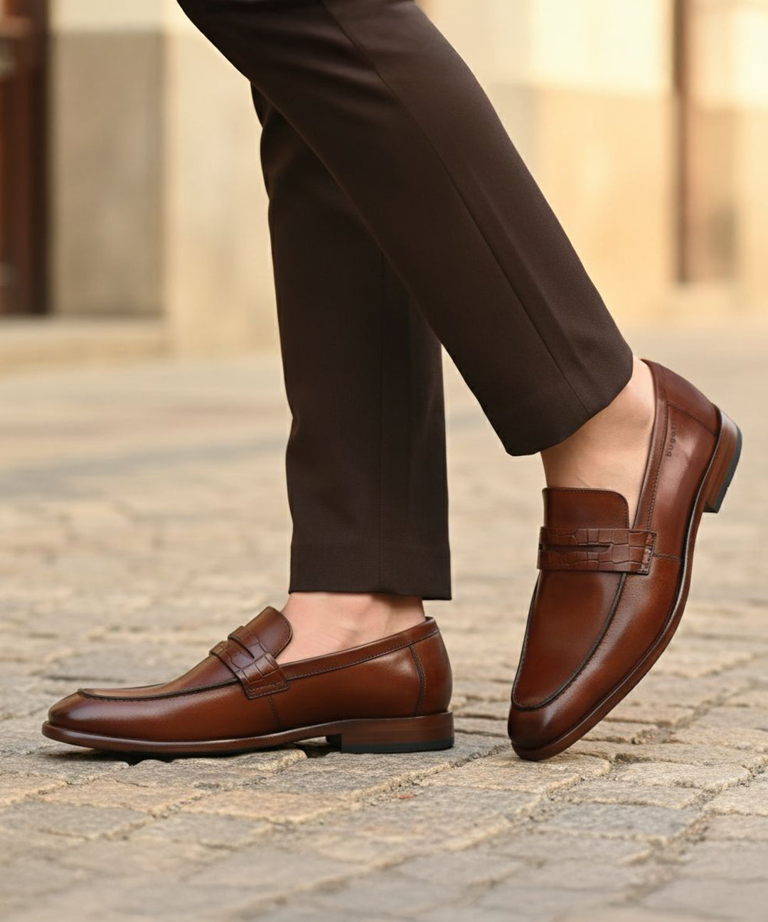 bugatti Mansueto cognac leather penny loafers with a textured strap detail, styled with brown trousers.
