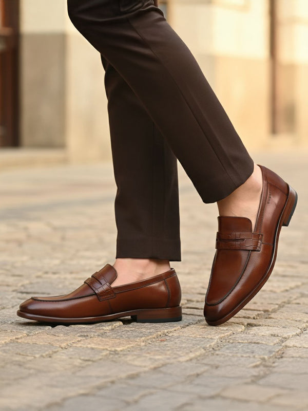 bugatti Mansueto cognac leather penny loafers with a textured strap detail, styled with brown trousers.
