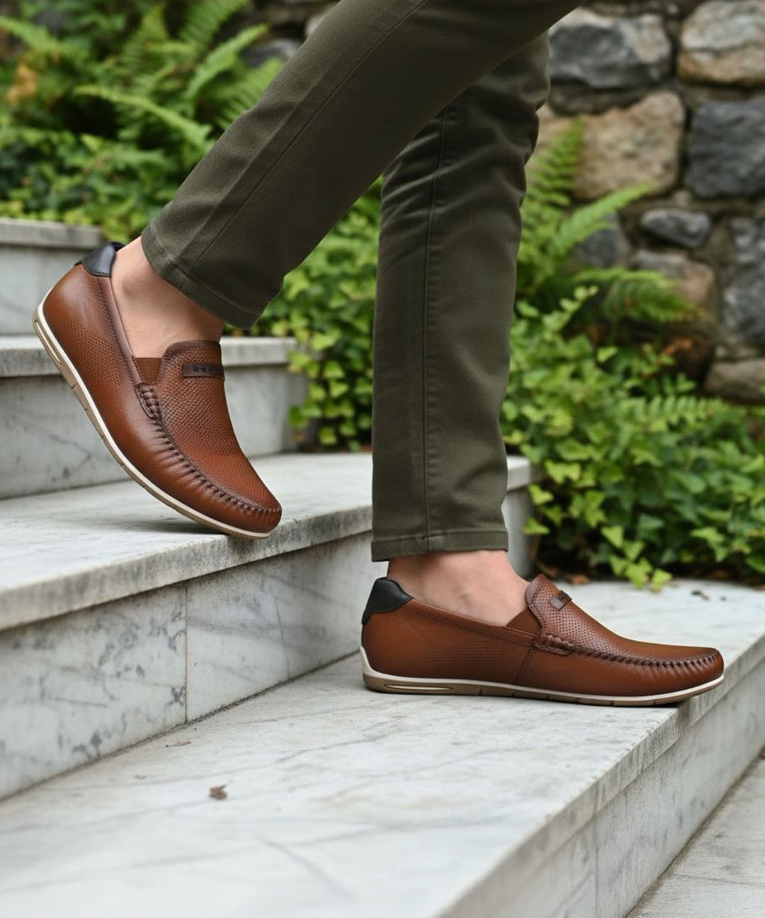 bugatti Chesley cognac leather slip-on shoe with a black accent at the heel, styled with olive pants on marble steps.
