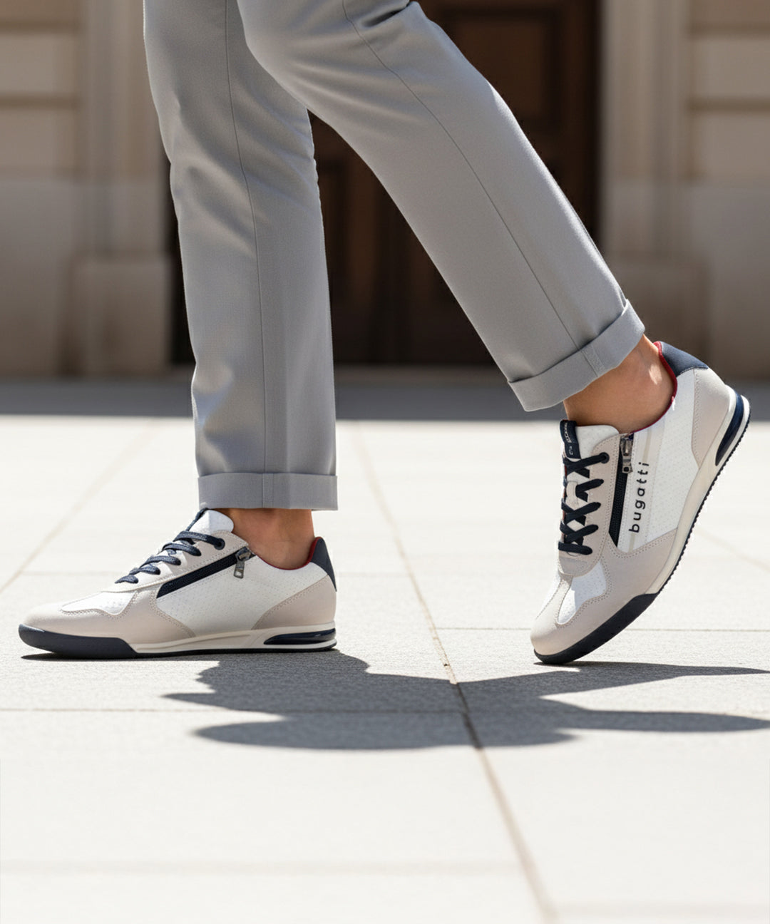Lifestyle shot of bugatti Trevor white sneakers, with zipper detail, worn with gray pants and walking on pavement.