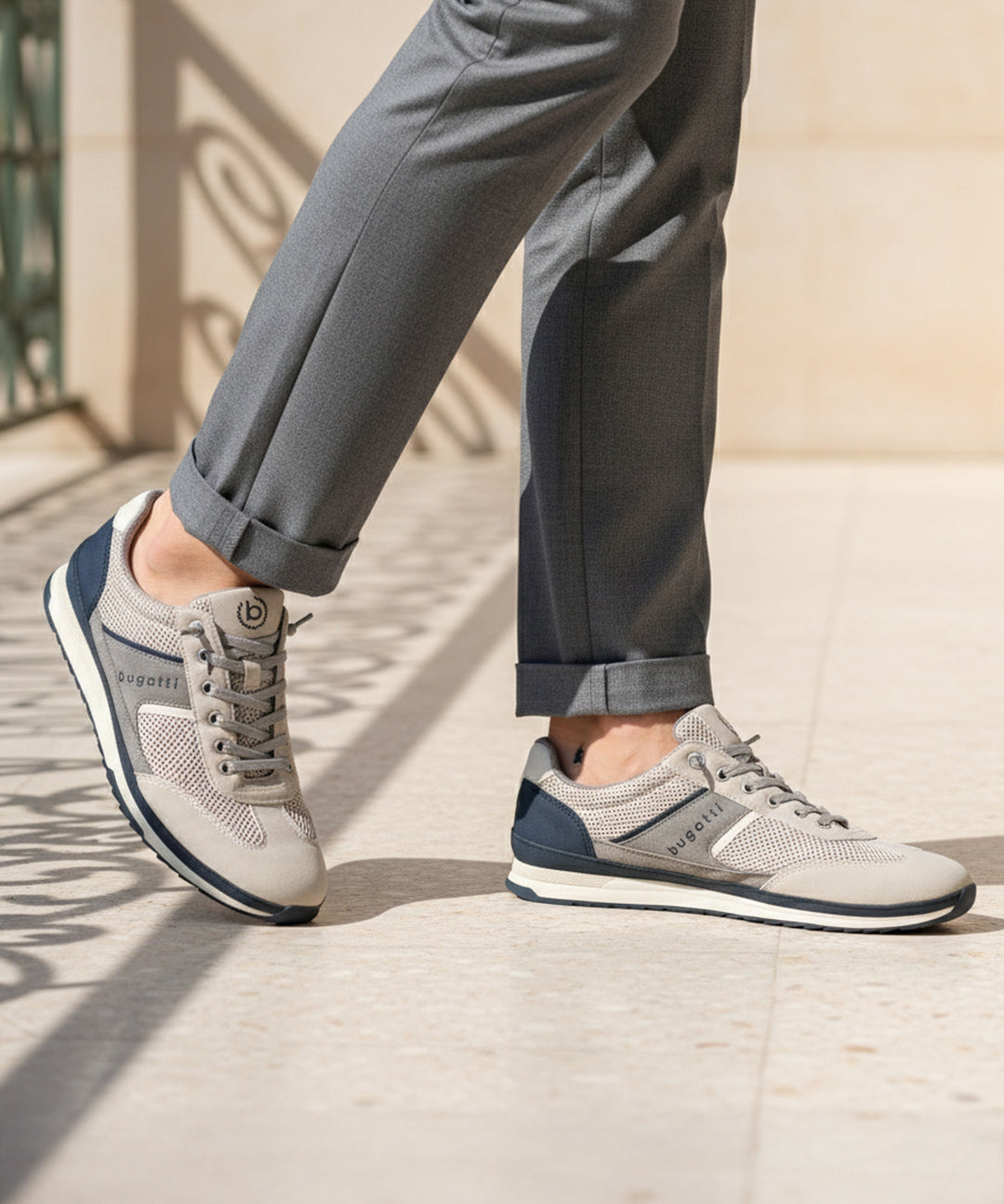 Lifestyle image of bugatti Riptide sand Comfort Sneakers, with gray mesh and suede upper, dark blue accents, paired with gray trousers.
