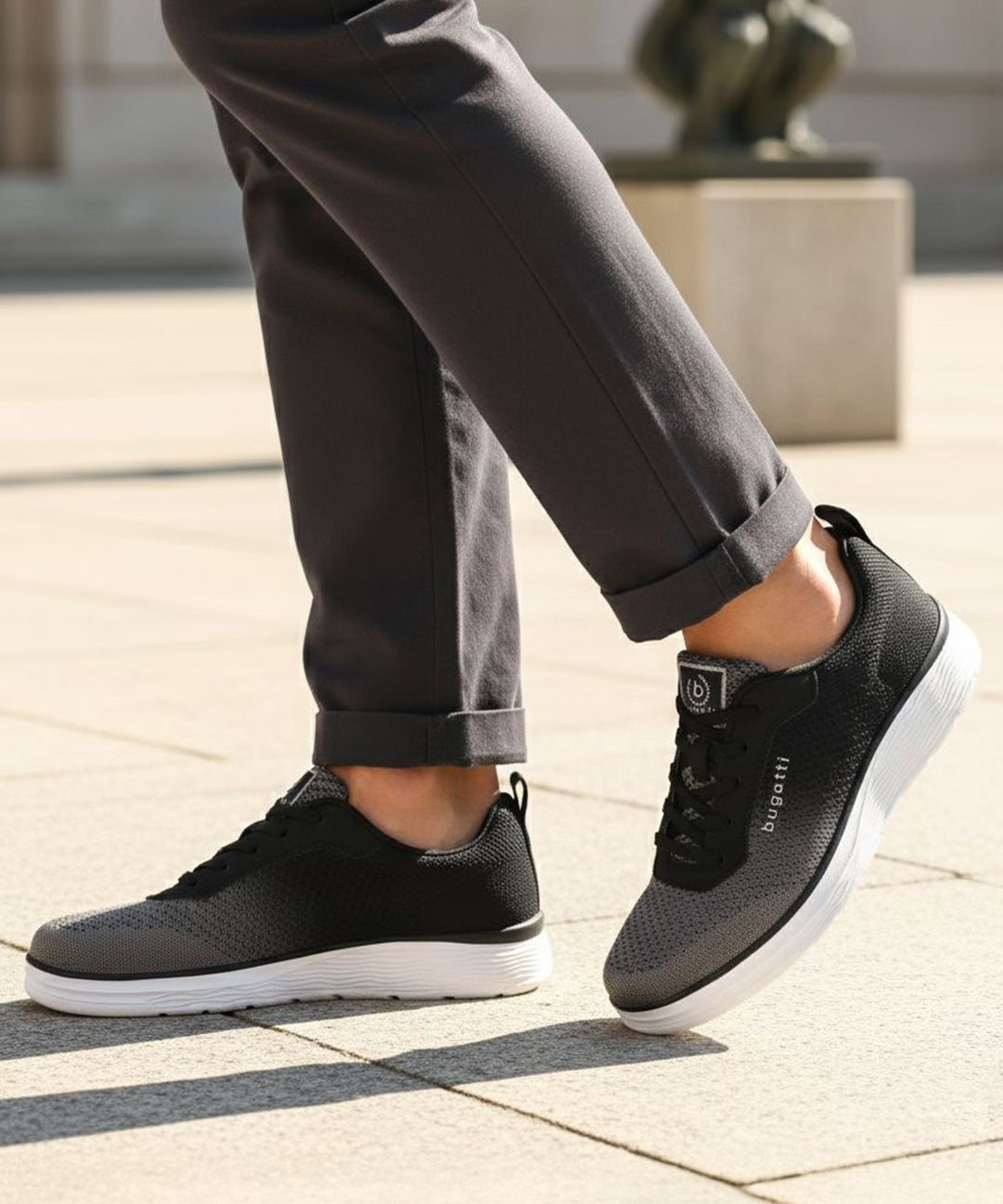 Lifestyle image of bugatti Astra athleisure sneakers in dark grey with a white sole, worn with rolled grey pants.
