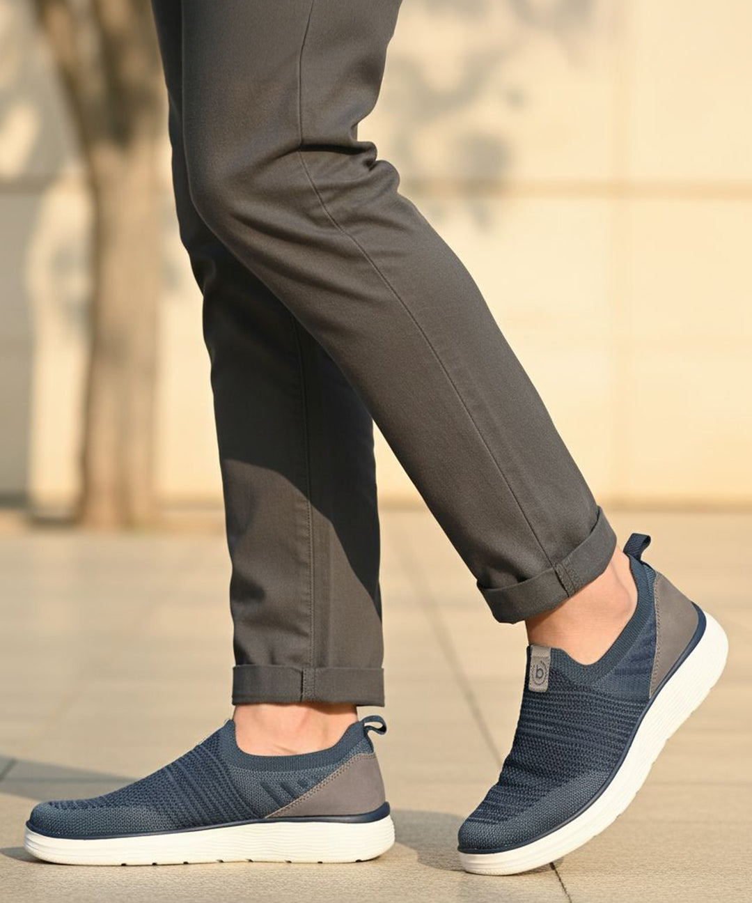 bugatti Astra blue slip-on casual shoes with a knit texture and white soles, styled with gray trousers.
