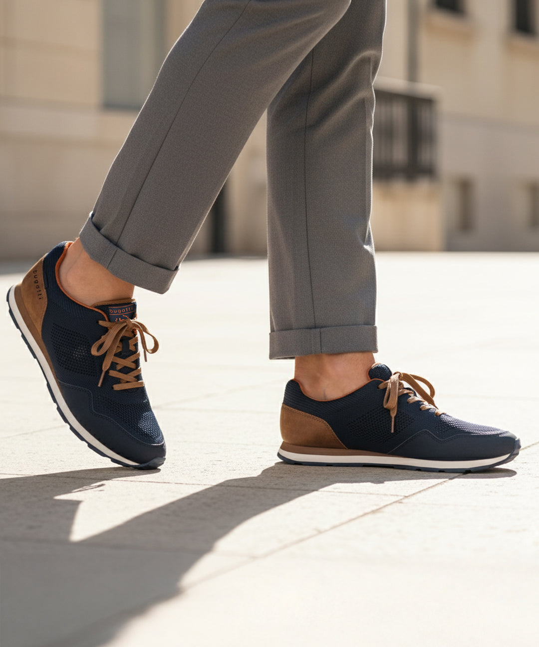 Lifestyle image of bugatti Sacramento dark blue sneakers with brown accents and laces, worn with cuffed grey trousers.
