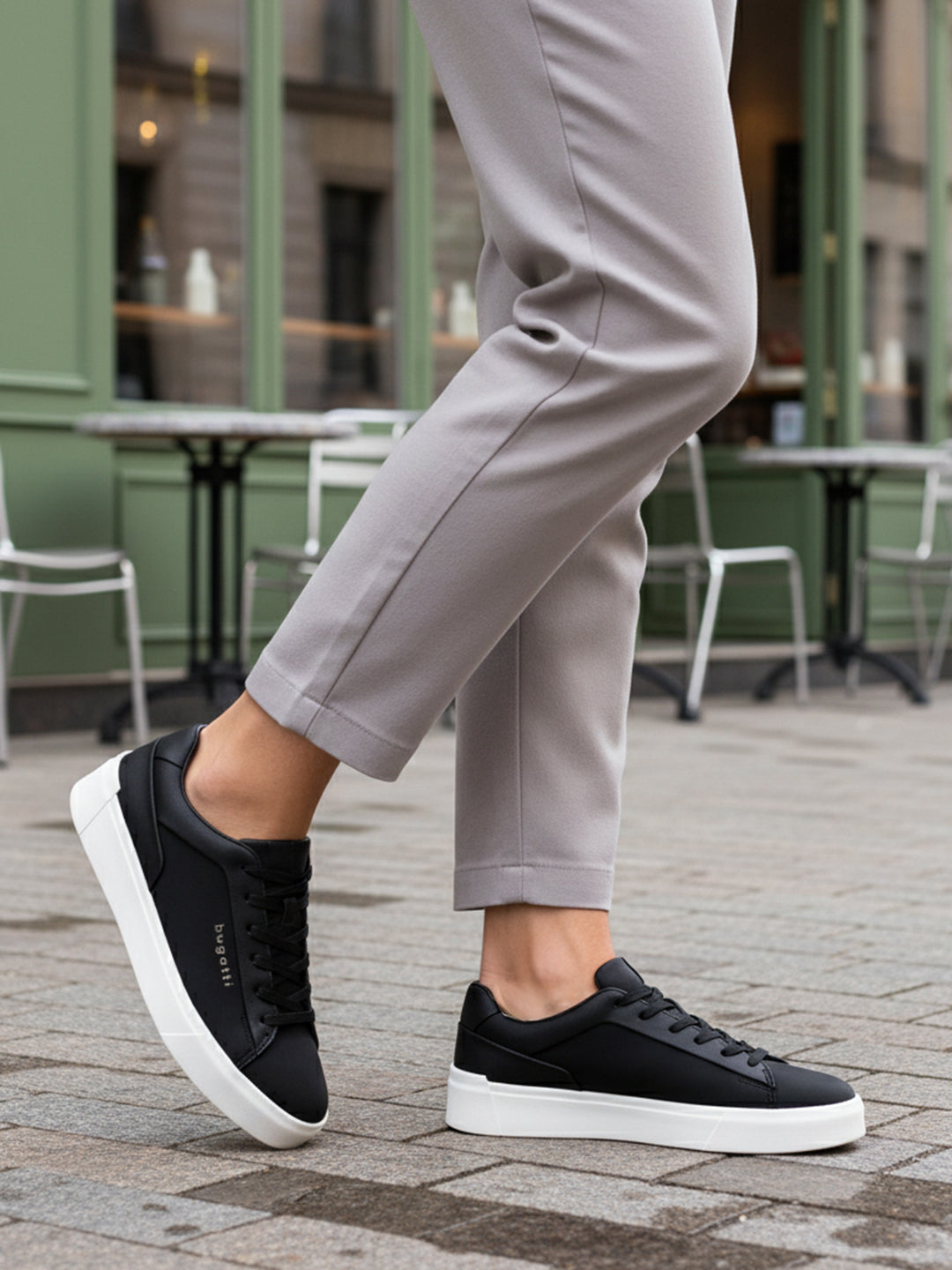 Lifestyle shot of bugatti Gunter black sneakers with a white sole, worn with grey pants.
