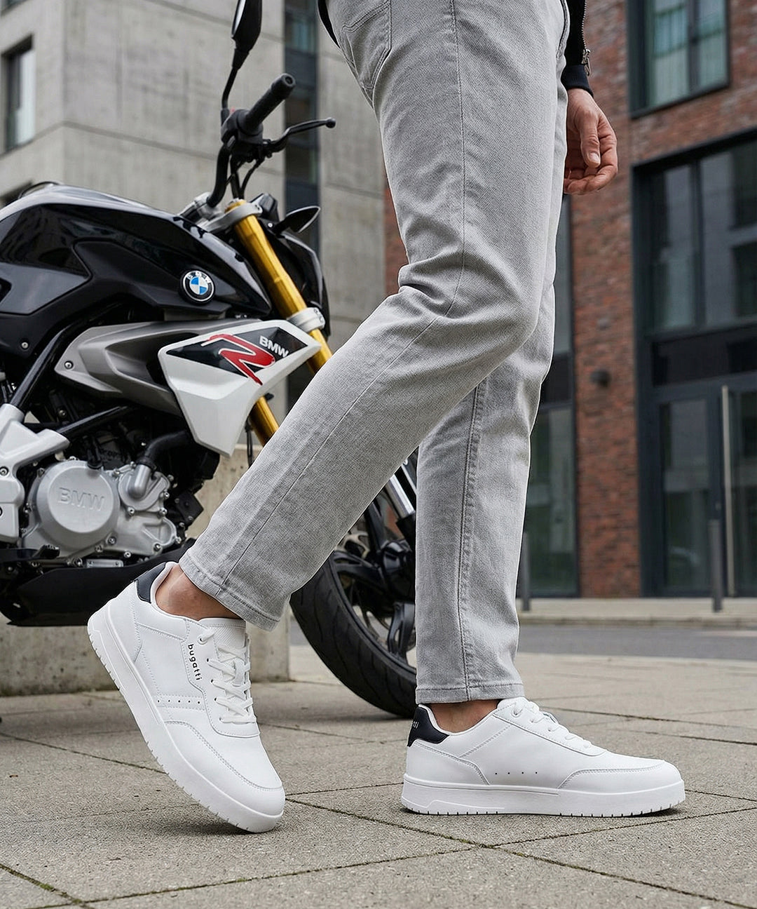 Lifestyle close-up of white bugatti Helium Comfort Sneakers, worn with grey pants and standing next to a motorcycle.
