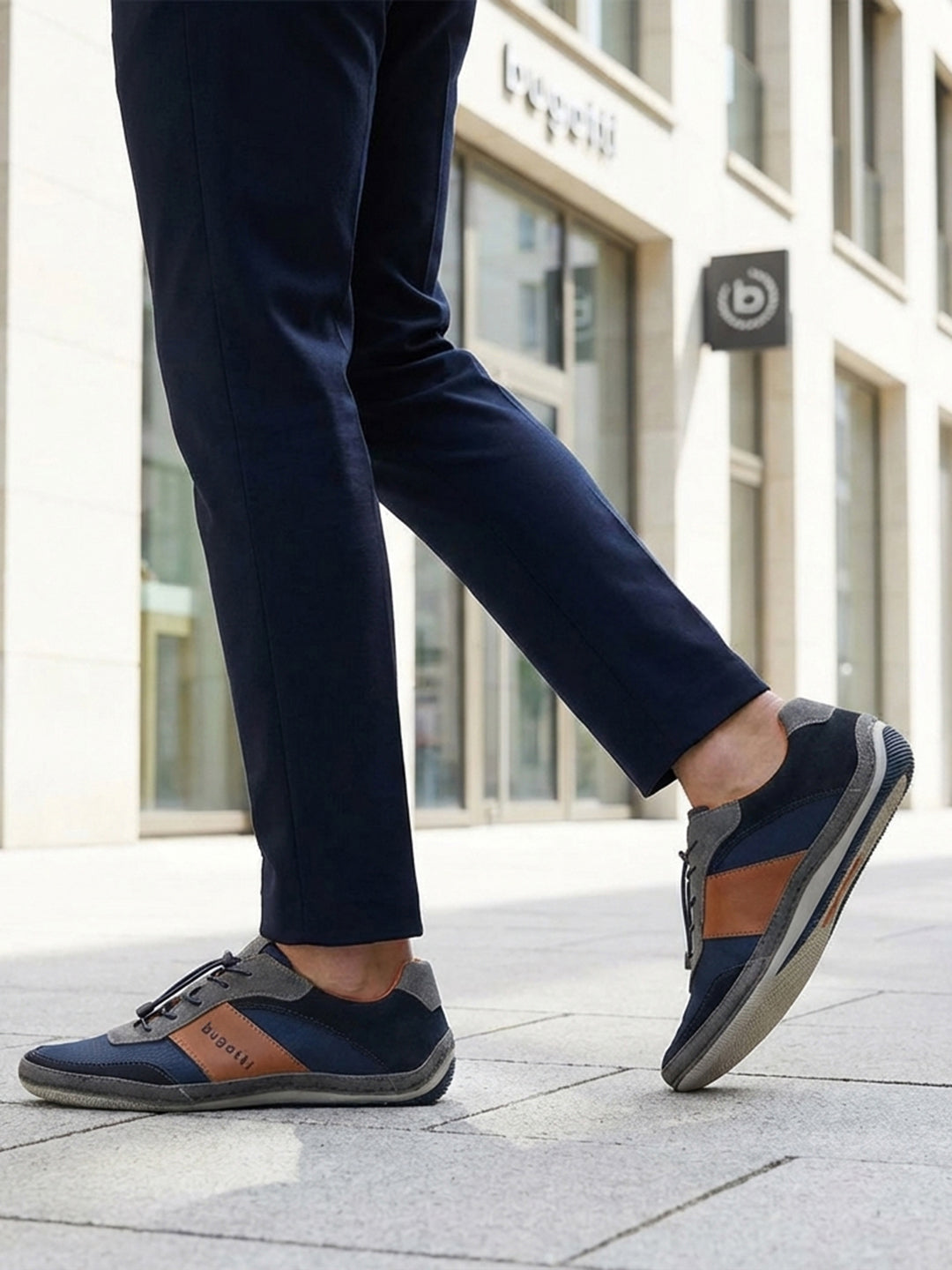 Lifestyle shot of a person wearing navy trousers and bugatti Canario Grey sneakers with brown leather accents.