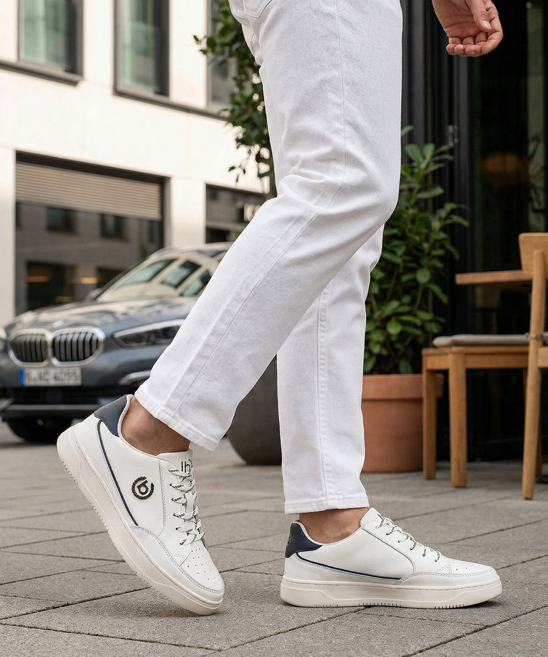 Lifestyle shot of bugatti Humus white premium leather mid-top sneakers with dark blue heel detail, worn with white pants.
