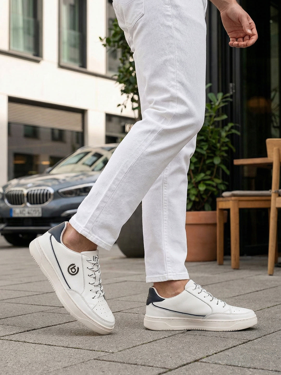 Lifestyle shot of bugatti Humus white premium leather mid-top sneakers with dark blue heel detail, worn with white pants.
