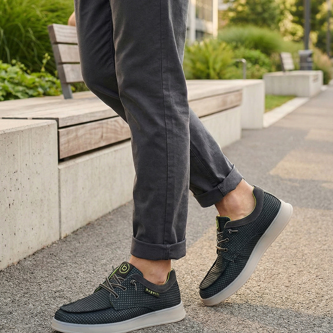Lifestyle view of bugatti Fiero black comfort sneakers, worn with rolled dark gray pants, on a paved walkway.
