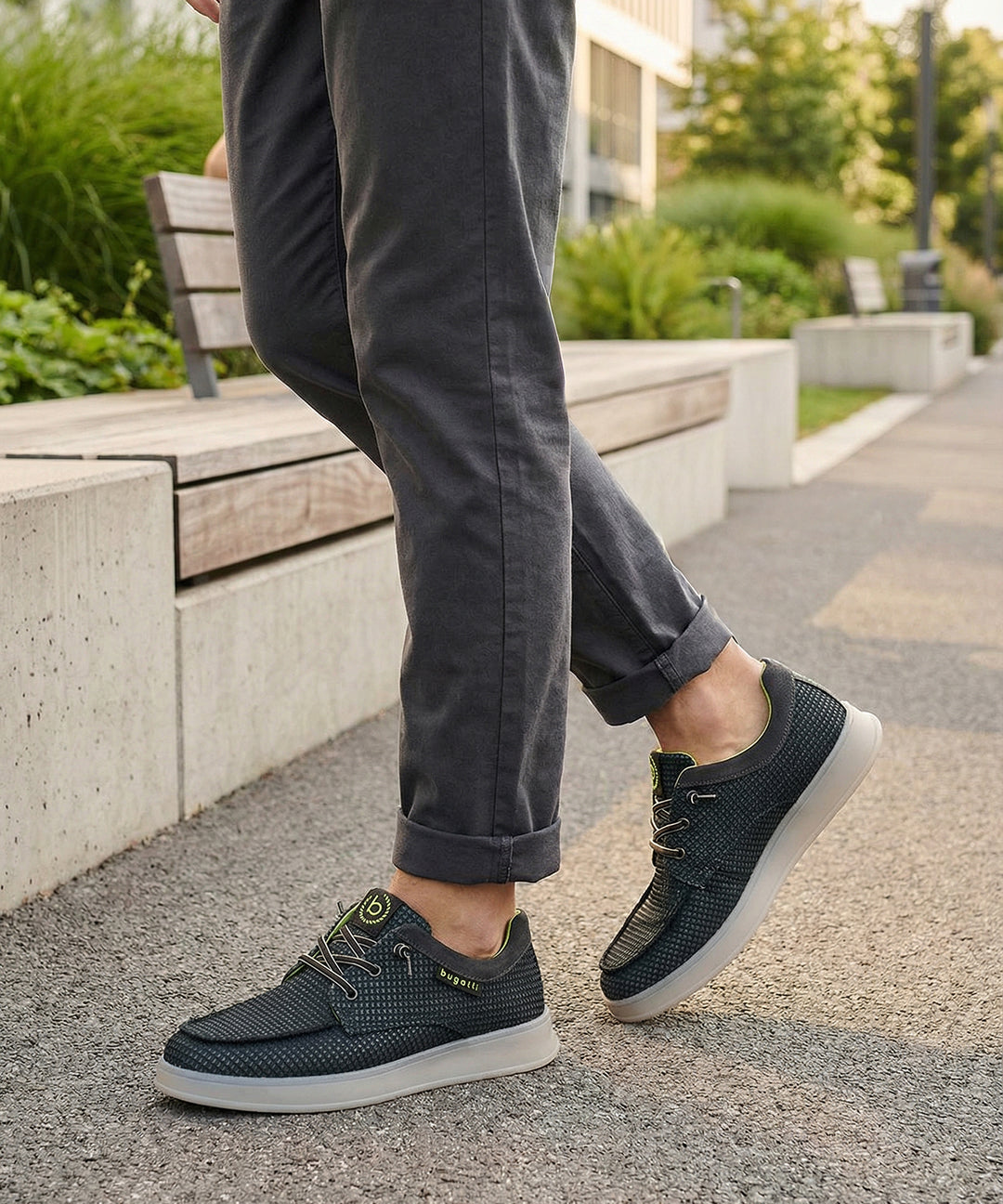Lifestyle view of bugatti Fiero black comfort sneakers, worn with rolled dark gray pants, on a paved walkway.

