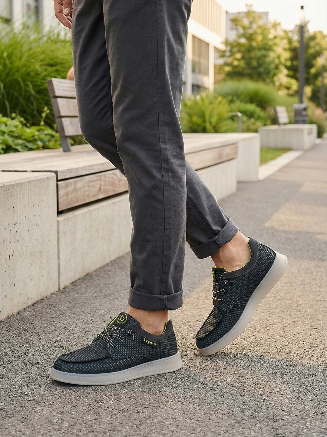 Lifestyle view of bugatti Fiero black comfort sneakers, worn with rolled dark gray pants, on a paved walkway.
