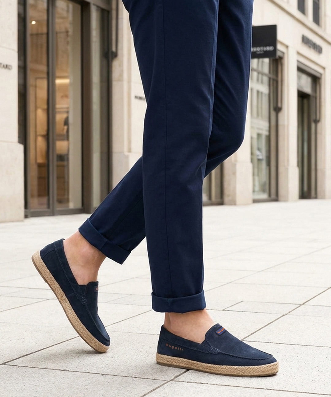 Lifestyle image of bugatti Spendril dark blue leather slip-on shoes with a jute sole, worn with cuffed navy pants.

