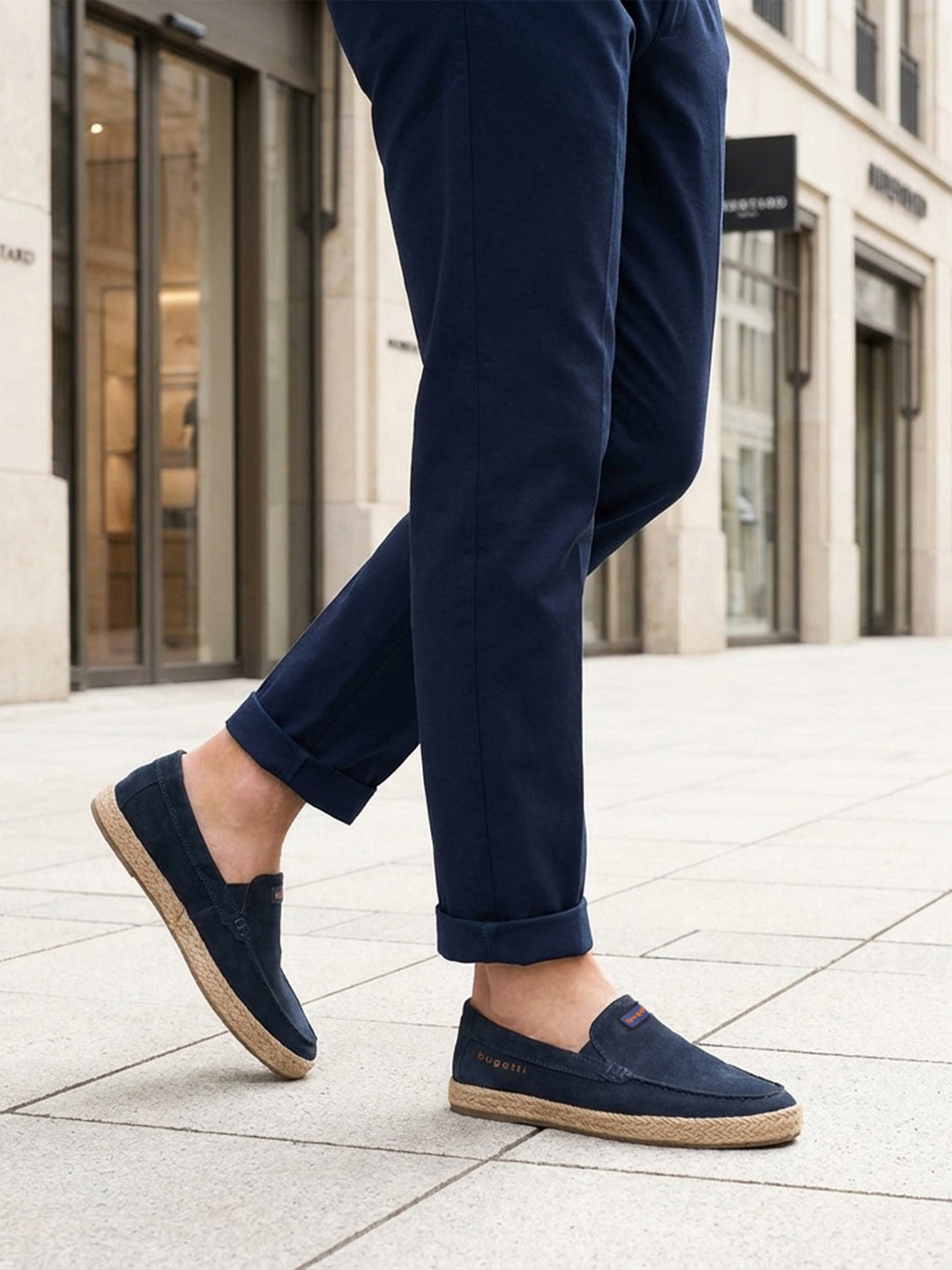 Lifestyle image of bugatti Spendril dark blue leather slip-on shoes with a jute sole, worn with cuffed navy pants.
