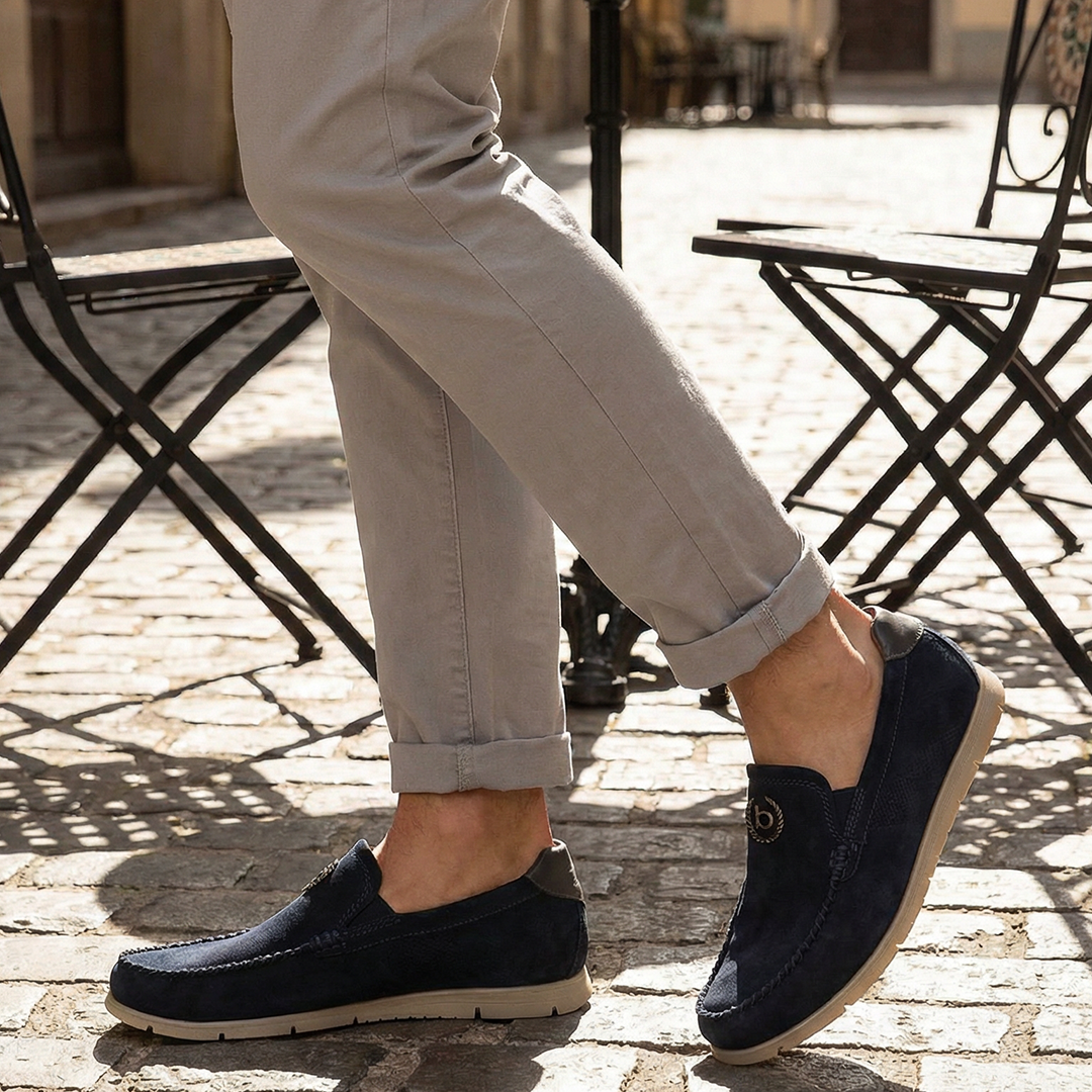 Lifestyle shot of bugatti Clipperton dark blue leather slip-on shoes with grey pants and iron chairs in the background.