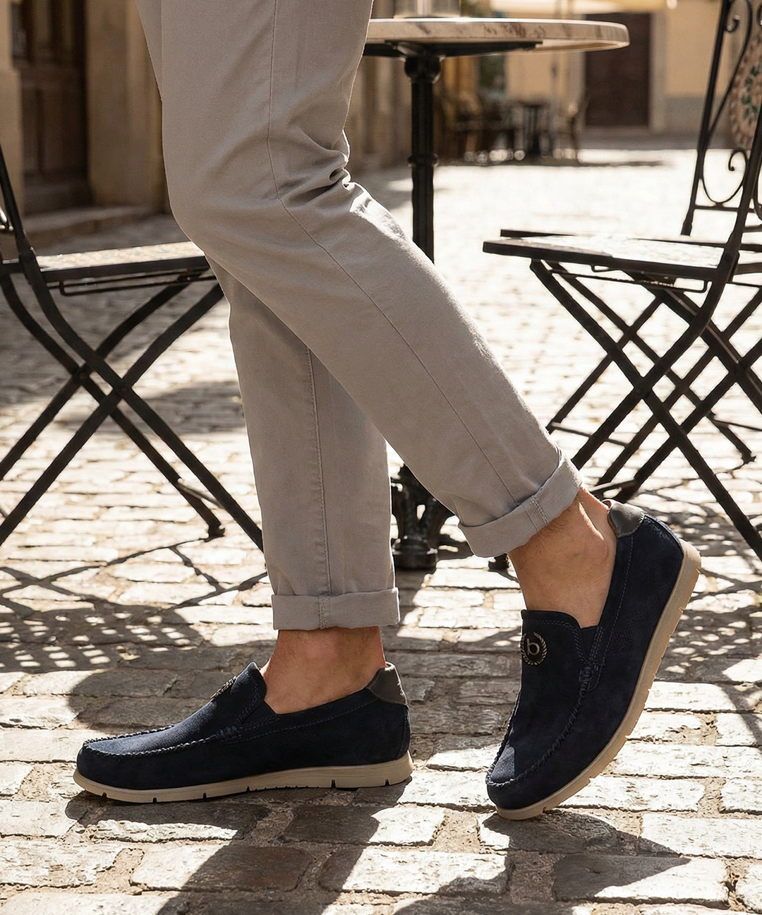 Lifestyle shot of bugatti Clipperton dark blue leather slip-on shoes with grey pants and iron chairs in the background.