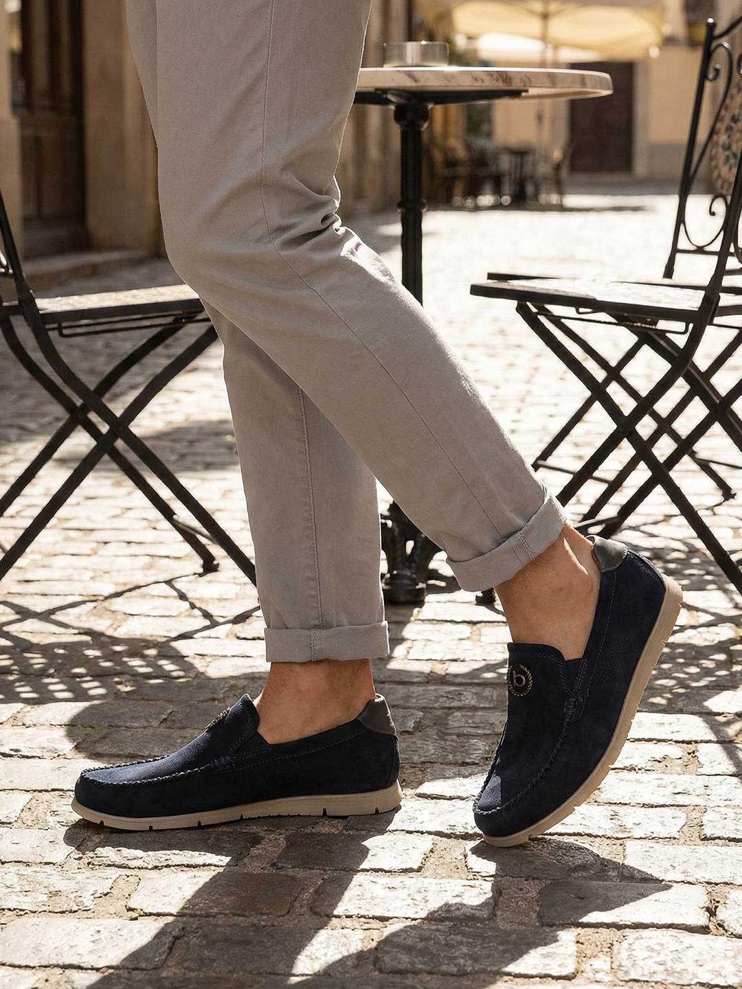 Lifestyle shot of bugatti Clipperton dark blue leather slip-on shoes with grey pants and iron chairs in the background.