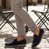 Lifestyle shot of bugatti Clipperton dark blue leather slip-on shoes with grey pants and iron chairs in the background.