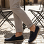 Lifestyle shot of bugatti Clipperton dark blue leather slip-on shoes with grey pants and iron chairs in the background.
