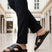 bugatti Cobie black leather sandals with a cross-strap design, paired with black trousers in a lifestyle shot.

