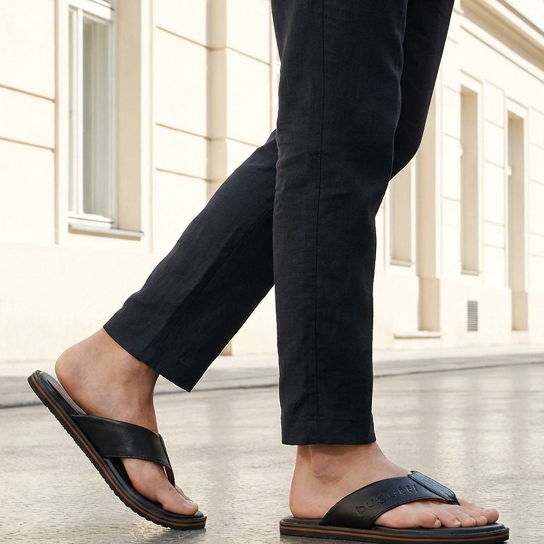 Lifestyle shot of black bugatti leather thong sandals with brown soles, worn with dark trousers.
