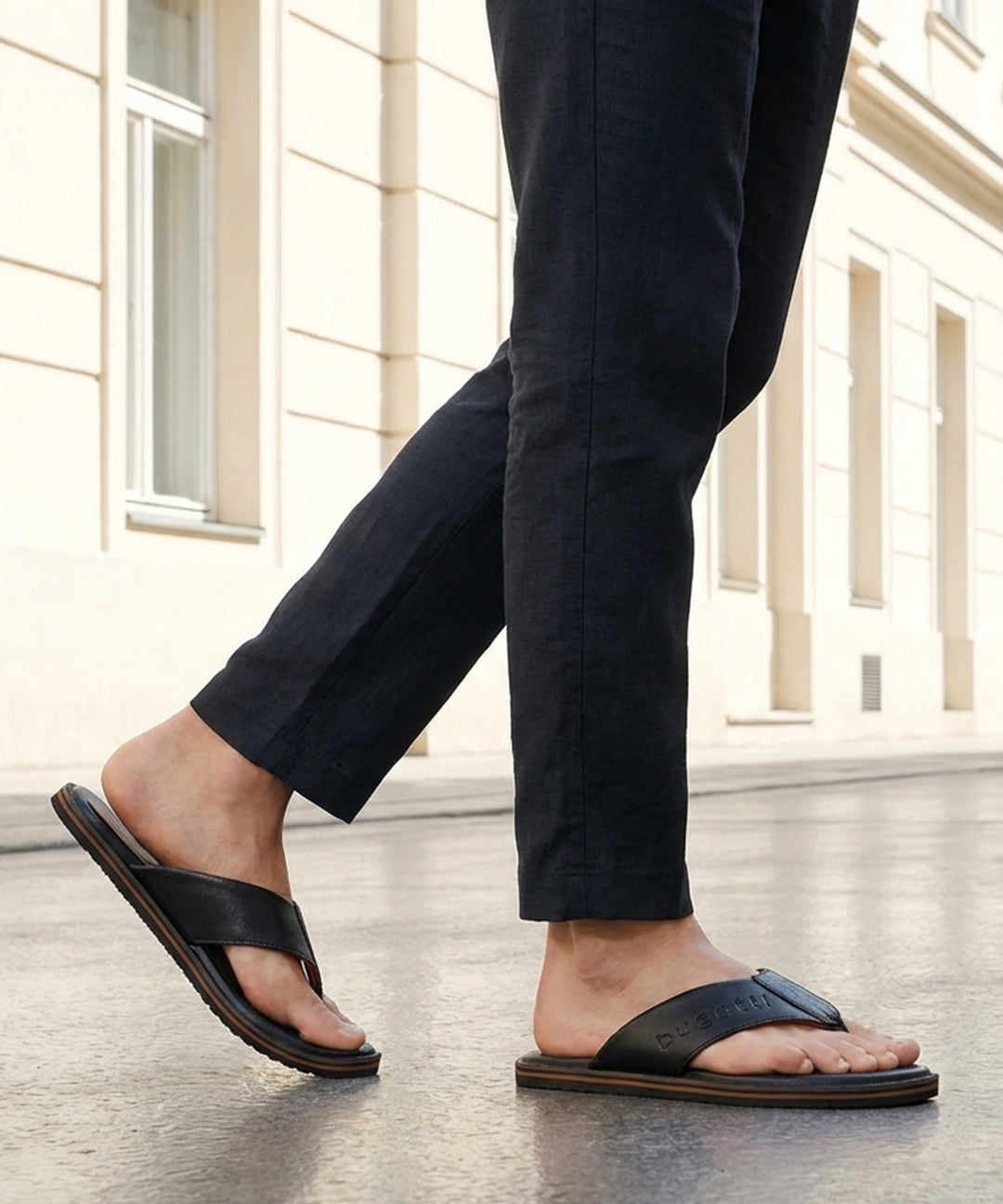 Lifestyle shot of black bugatti leather thong sandals with brown soles, worn with dark trousers.
