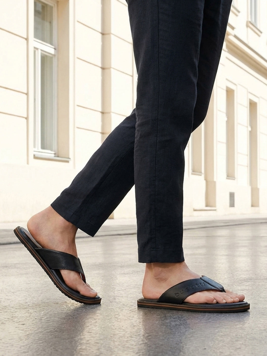 Lifestyle shot of black bugatti leather thong sandals with brown soles, worn with dark trousers.

