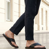 Lifestyle shot of black bugatti leather thong sandals with brown soles, worn with dark trousers.
