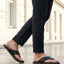 Lifestyle shot of black bugatti leather thong sandals with brown soles, worn with dark trousers.
