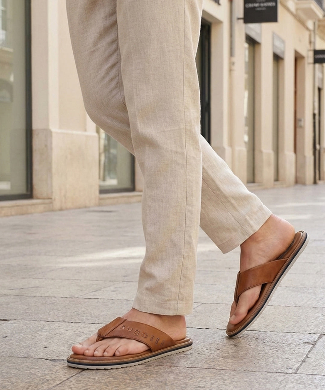 Lifestyle image of bugatti Cobie cognac leather thong sandals worn with casual tan pants in a city setting.